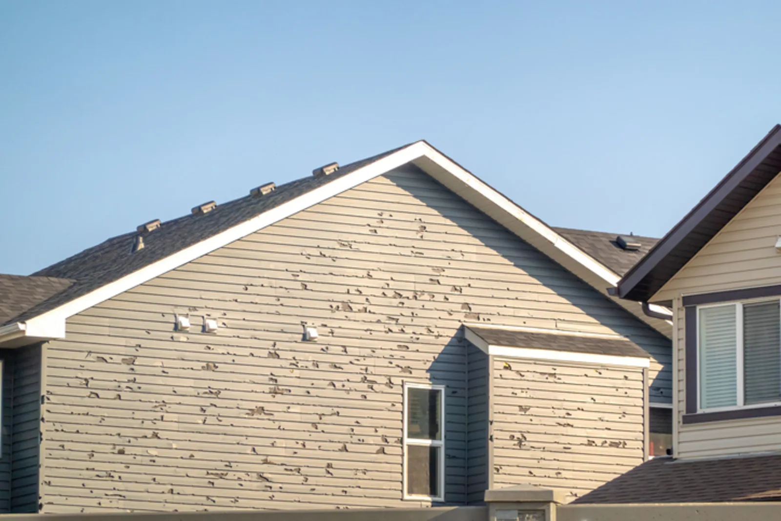 Tan vinyl siding on a suburban home cracked and punched with holes across the gable elevation by wind-driven hail