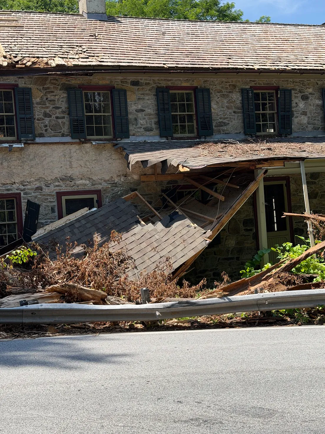 Storm-damaged Pennsylvania stone farmhouse with a collapsed porch roof and downed tree debris in the front yard